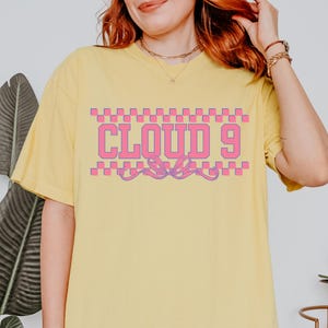 Puede incluir: Camiseta amarilla clara con el texto "CLOUD 9" en rosa, contorneado en blanco, y un patr&oacute;n de cuadros arriba y abajo. La camiseta tiene mangas cortas y un corte holgado.