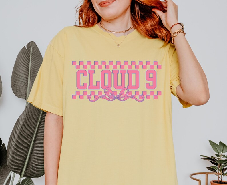 Puede incluir: Camiseta amarilla clara con el texto "CLOUD 9" en rosa, contorneado en blanco, y un patr&oacute;n de cuadros arriba y abajo. La camiseta tiene mangas cortas y un corte holgado.