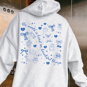 May include: A light gray hoodie with a hood and long sleeves. The back of the hoodie features a blue graphic design with the text "the world is hard on beautiful things" and various illustrations of bows, hearts, and stars.