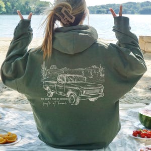 May include: An olive green hooded sweatshirt featuring a white illustration of a classic pickup truck and desert scenery. The text reads "You won't find no answers safe at home". The hoodie is worn outside.