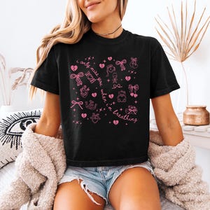 Pode incluir: Camiseta preta com ilustra&ccedil;&otilde;es gr&aacute;ficas rosa de la&ccedil;os, cora&ccedil;&otilde;es e outros s&iacute;mbolos. O texto "I only miss you when I'm breathing" &eacute; impresso em rosa. A camisa &eacute; de manga curta e tem gola redonda.