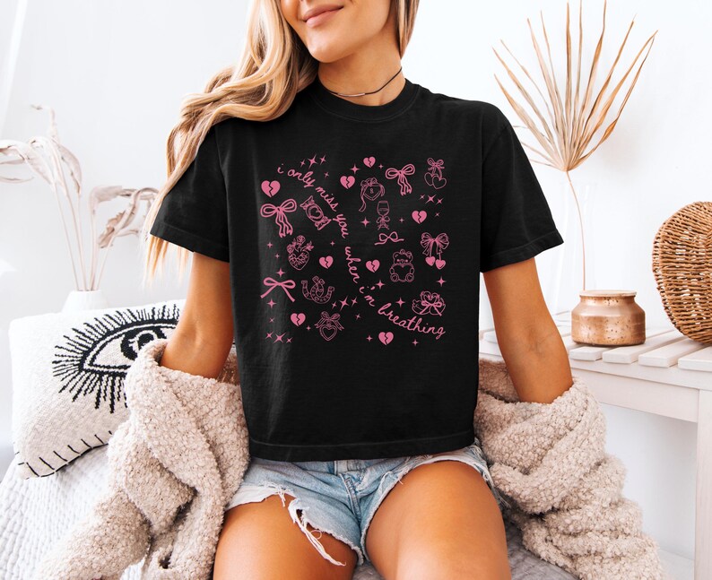 Pode incluir: Camiseta preta com ilustra&ccedil;&otilde;es gr&aacute;ficas rosa de la&ccedil;os, cora&ccedil;&otilde;es e outros s&iacute;mbolos. O texto "I only miss you when I'm breathing" &eacute; impresso em rosa. A camisa &eacute; de manga curta e tem gola redonda.