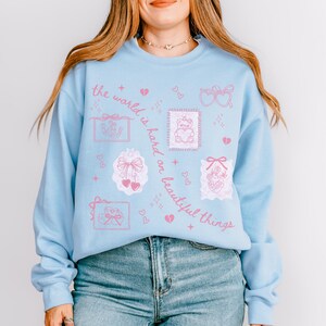 K&ouml;nnte beinhalten: Hellblaues Sweatshirt mit rosa Grafikdesign. Das Design umfasst Herzen, Schleifen und den Satz "die Welt ist hart zu sch&ouml;nen Dingen". Das lang&auml;rmlige Oberteil ist aus einem weichen Material gefertigt.
