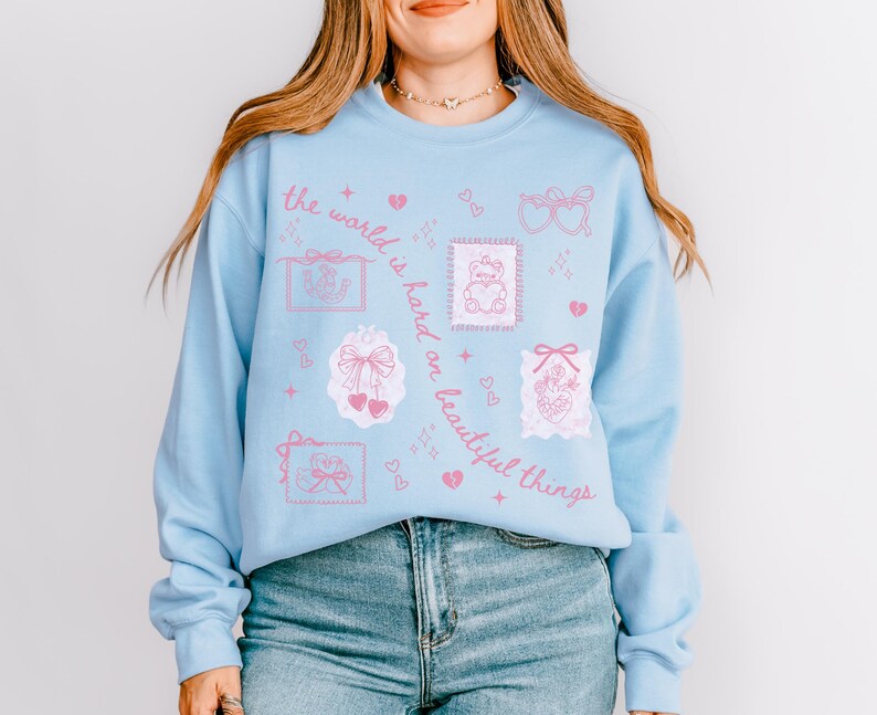 K&ouml;nnte beinhalten: Hellblaues Sweatshirt mit rosa Grafikdesign. Das Design umfasst Herzen, Schleifen und den Satz "die Welt ist hart zu sch&ouml;nen Dingen". Das lang&auml;rmlige Oberteil ist aus einem weichen Material gefertigt.
