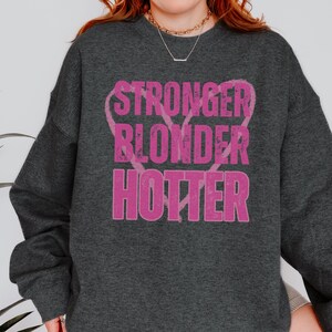 Puede incluir: Sudadera gris oscuro con las palabras "STRONGER BLONDER HOTTER" en letras rosas desgastadas. El texto est&aacute; dispuesto verticalmente con una forma de coraz&oacute;n detr&aacute;s de la palabra "BLONDER". La sudadera tiene mangas largas y cuello redondo.