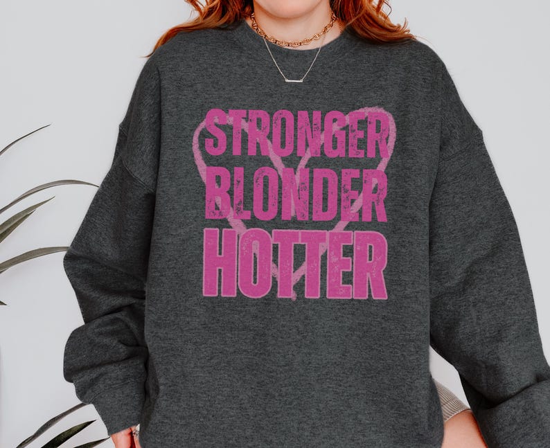 Puede incluir: Sudadera gris oscuro con las palabras "STRONGER BLONDER HOTTER" en letras rosas desgastadas. El texto est&aacute; dispuesto verticalmente con una forma de coraz&oacute;n detr&aacute;s de la palabra "BLONDER". La sudadera tiene mangas largas y cuello redondo.