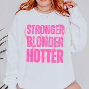 Puede incluir: Sudadera blanca con las palabras "STRONGER BLONDER HOTTER" en rosa brillante, con forma de coraz&oacute;n rosa detr&aacute;s de las palabras. La sudadera tiene mangas largas y cuello redondo.