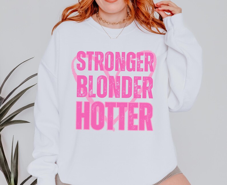 Puede incluir: Sudadera blanca con las palabras "STRONGER BLONDER HOTTER" en rosa brillante, con forma de coraz&oacute;n rosa detr&aacute;s de las palabras. La sudadera tiene mangas largas y cuello redondo.