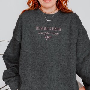 Może przedstawiać: Ciemnoszara bluza z r&oacute;żowym napisem "THE WORLD IS HARD ON beautiful things" i r&oacute;żową kokardą. Bluza z długim rękawem ma okrągły dekolt.