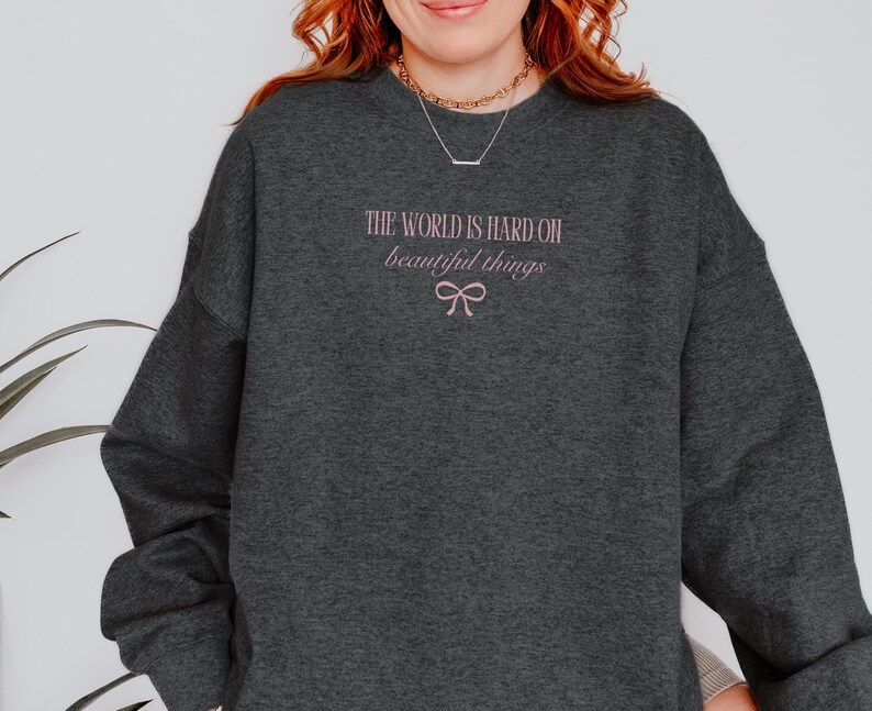 Może przedstawiać: Ciemnoszara bluza z r&oacute;żowym napisem "THE WORLD IS HARD ON beautiful things" i r&oacute;żową kokardą. Bluza z długim rękawem ma okrągły dekolt.