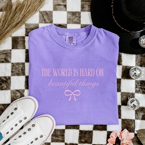 Puede incluir: Una camiseta lavanda con el texto rosa "THE WORLD IS HARD ON beautiful things" y un lazo. La camiseta est&aacute; doblada sobre una superficie a cuadros, con zapatillas blancas, un sombrero negro y adornos de bola de discoteca.