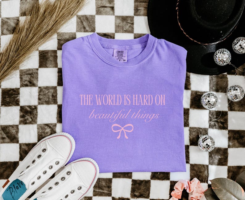 Puede incluir: Una camiseta lavanda con el texto rosa "THE WORLD IS HARD ON beautiful things" y un lazo. La camiseta est&aacute; doblada sobre una superficie a cuadros, con zapatillas blancas, un sombrero negro y adornos de bola de discoteca.