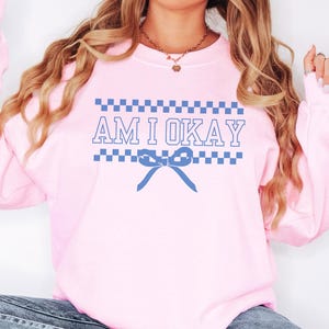 May include: A light pink sweatshirt with the text "AM I OKAY" in blue. The text is framed by blue and white checkered patterns, with a blue bow graphic below the text. The sweatshirt is a casual style.
