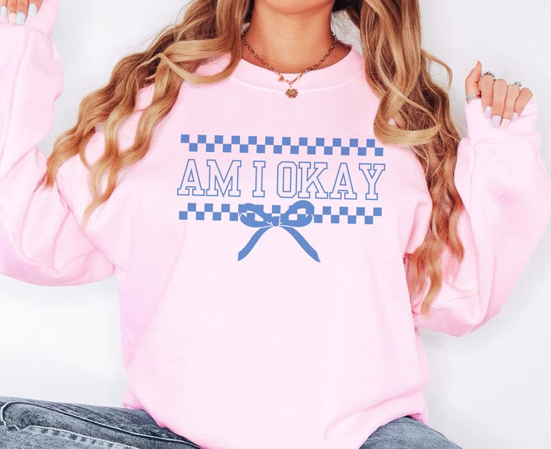 May include: A light pink sweatshirt with the text "AM I OKAY" in blue. The text is framed by blue and white checkered patterns, with a blue bow graphic below the text. The sweatshirt is a casual style.