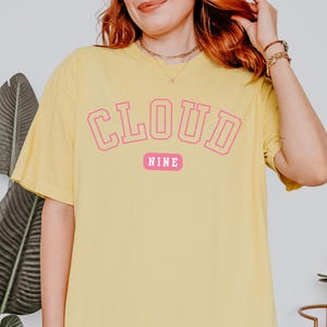May include: Yellow t-shirt with the text "CLOUD NINE" in pink block letters. The word "NINE" is on a pink rectangular background. The shirt has short sleeves and a relaxed fit.