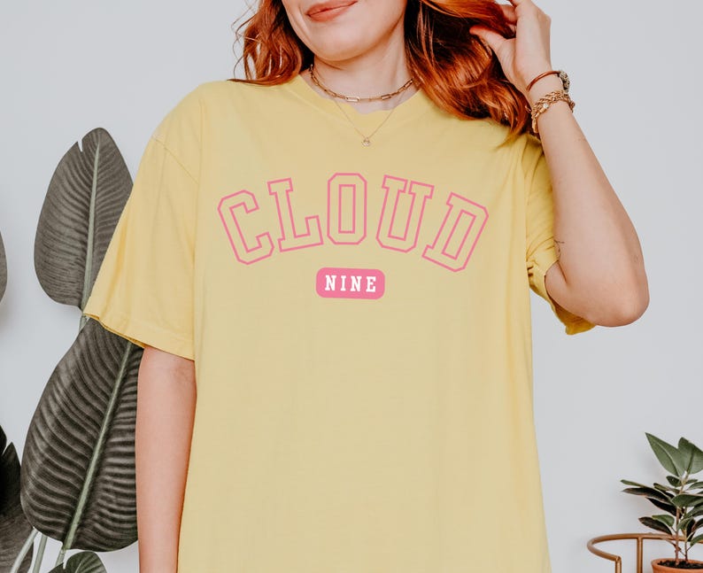 May include: Yellow t-shirt with the text "CLOUD NINE" in pink block letters. The word "NINE" is on a pink rectangular background. The shirt has short sleeves and a relaxed fit.