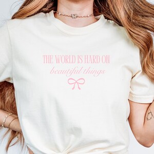 Puede incluir: Camiseta color crema con el texto rosa "THE WORLD IS HARD ON beautiful things" y un lazo rosa. La camiseta es de material suave y tiene mangas cortas.