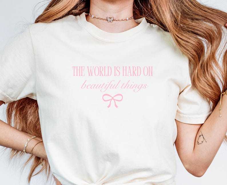 Puede incluir: Camiseta color crema con el texto rosa "THE WORLD IS HARD ON beautiful things" y un lazo rosa. La camiseta es de material suave y tiene mangas cortas.