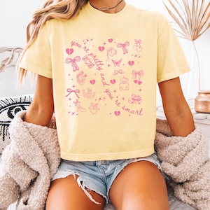 May include: A pale yellow short-sleeve t-shirt featuring a pink graphic print of various small icons including bows, hearts, a butterfly, a teddy bear, and a high-heeled shoe, with the text "you're pretty and you're smart" written diagonally.