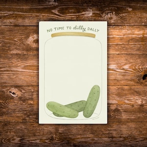 May include: A rectangular notepad with a jar illustration and the text "NO TIME TO dilly DALLY". The notepad features three green pickles at the bottom. The background is a light cream color.