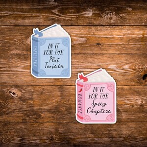 May include: Two book-shaped stickers on a wooden surface. One is light blue with the text "IN IT FOR THE Plot Twists" and "THRILLERS". The other is pink with "IN IT FOR THE Spicy Chapters" and "ROMANCE".