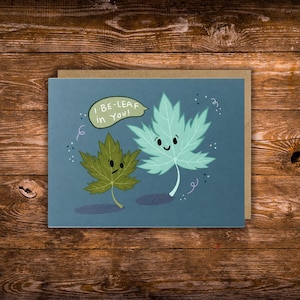 May include: Greeting card with a blue background featuring two cartoon leaves. One leaf is green, the other is teal. The text "I BE-LEAF in YOU!" is in a speech bubble above the leaves. The card is on a brown envelope.