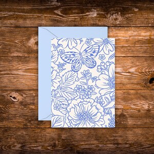May include: A greeting card with a light blue envelope. The card features a cream background with a blue floral and butterfly design. The card is placed on a wooden surface.