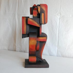 Op de afbeelding: Een verticaal houten beeldhouwwerk bestaande uit geometrische blokken en hoekige vormen. Het object heeft een kleurverloop van diepzwart, gebrand oranje en helderrood, met een gladde afwerking op een rechthoekige voet in een rechtopstaande positie.