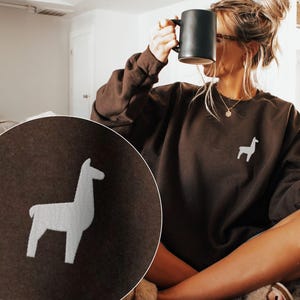May include: A brown sweatshirt with a white llama graphic. The sweatshirt is long-sleeved and has a crew neck. A black mug is held up near the face. The person is sitting on a couch.