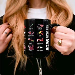 Peut inclure: Mug en c&eacute;ramique noire avec l'inscription "2026 Grand Prix Formula One Calendar" et des trac&eacute;s de circuits de course avec des drapeaux. Les lieux incluent Djeddah, Miami et Silverstone. Le mug est tenu par une personne.