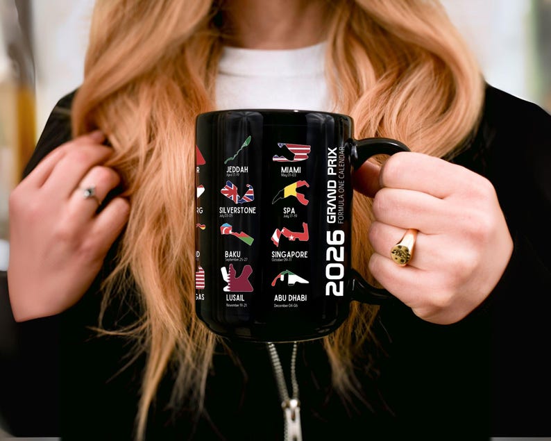 Peut inclure: Mug en c&eacute;ramique noire avec l'inscription "2026 Grand Prix Formula One Calendar" et des trac&eacute;s de circuits de course avec des drapeaux. Les lieux incluent Djeddah, Miami et Silverstone. Le mug est tenu par une personne.