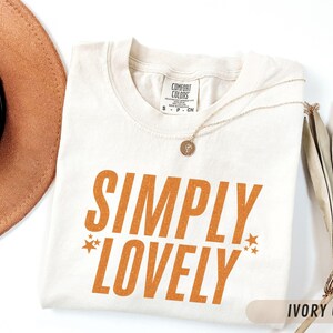 May include: Cream-coloured t-shirt with the words "SIMPLY LOVELY" in orange, with star accents. A gold necklace with a pendant lies on the shirt. A brown hat and dried plants are in the background.