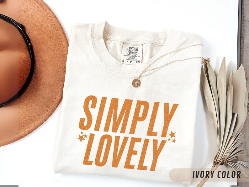 May include: Cream-coloured t-shirt with the words "SIMPLY LOVELY" in orange, with star accents. A gold necklace with a pendant lies on the shirt. A brown hat and dried plants are in the background.