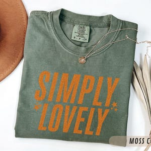 May include: A moss green t-shirt with the words "SIMPLY LOVELY" in orange block letters. The shirt has a crew neck and short sleeves. A gold necklace and a decorative dried plant are also in the image.