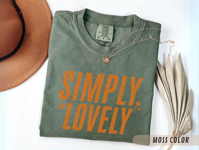 May include: A moss green t-shirt with the words "SIMPLY LOVELY" in orange block letters. The shirt has a crew neck and short sleeves. A gold necklace and a decorative dried plant are also in the image.