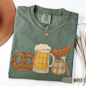 May include: Moss green t-shirt with a graphic of a pretzel, beer stein, sausage, and a plaid pumpkin. The shirt has a crew neck and short sleeves. A silver necklace with a pendant is on the shirt. The words "MOSS COLOR" are at the bottom right.