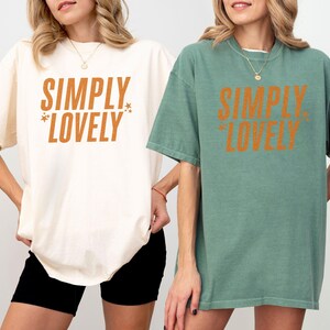 May include: Two oversized t-shirts, one ivory and one light green, each with the text "SIMPLY LOVELY" in orange. The ivory shirt is paired with black shorts. The light green shirt is paired with a necklace.