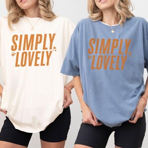 May include: Two oversized t-shirts, one cream and one blue, each with the text "SIMPLY LOVELY" in orange. The shirts are paired with black shorts. The image showcases casual, comfortable apparel.