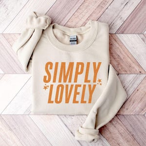 Pu&ograve; includere: Felpa color crema con le parole "SIMPLY LOVELY" in lettere arancioni. Due piccole grafiche a stella affiancano la parola "LOVELY". La felpa &egrave; piegata su una superficie di legno.