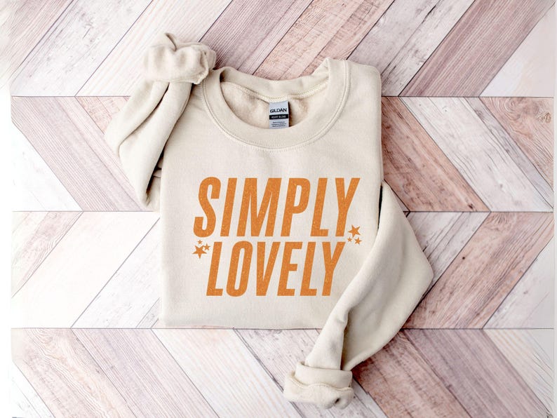 Pu&ograve; includere: Felpa color crema con le parole "SIMPLY LOVELY" in lettere arancioni. Due piccole grafiche a stella affiancano la parola "LOVELY". La felpa &egrave; piegata su una superficie di legno.