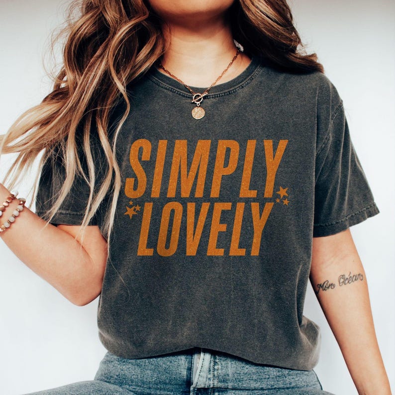 Vintage black Comfort Colors® t-shirt with bold mustard text that says “Simply Lovely” and small star graphics, worn by a woman with wavy hair.