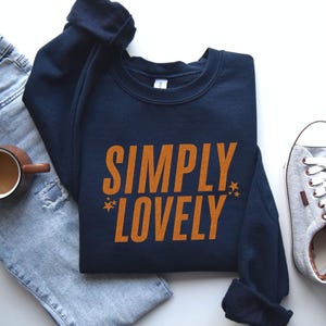 Pu&ograve; includere: Felpa blu navy con la scritta "SIMPLY LOVELY" in arancione, con due piccole stelle. La felpa &egrave; accanto a un paio di jeans azzurri, una tazza di caff&egrave; e scarpe da ginnastica grigie.