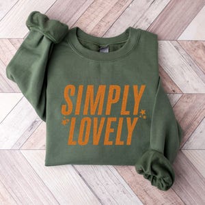 Pu&ograve; includere: Una felpa verde oliva con la scritta "SIMPLY LOVELY" stampata in arancione. Il testo &egrave; centrato sulla parte anteriore del capo, con piccoli grafici a stella su entrambi i lati della parola "LOVELY". Le maniche sono arricciate.
