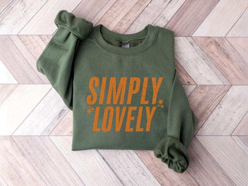 Pu&ograve; includere: Una felpa verde oliva con la scritta "SIMPLY LOVELY" stampata in arancione. Il testo &egrave; centrato sulla parte anteriore del capo, con piccoli grafici a stella su entrambi i lati della parola "LOVELY". Le maniche sono arricciate.
