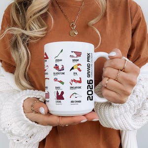 Peut inclure: Mug en c&eacute;ramique blanche avec le calendrier de Formule 1 2026, affichant des lieux de course comme Miami, Silverstone et Abu Dhabi. La tasse a une anse et est tenue par une personne.
