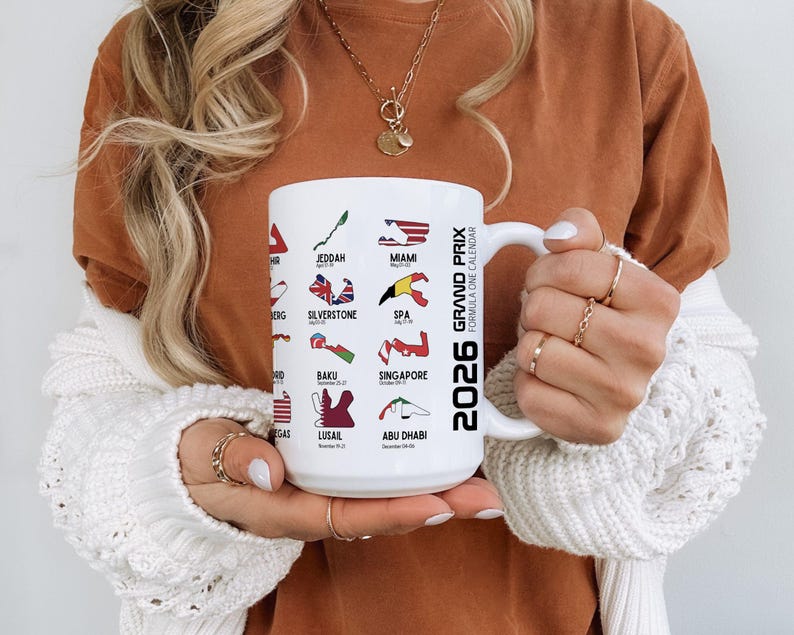 Peut inclure: Mug en c&eacute;ramique blanche avec le calendrier de Formule 1 2026, affichant des lieux de course comme Miami, Silverstone et Abu Dhabi. La tasse a une anse et est tenue par une personne.
