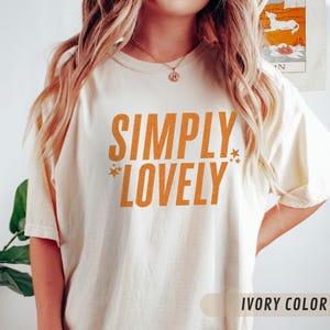 May include: Cream-coloured t-shirt featuring the orange text "SIMPLY LOVELY". The design includes star graphics. The shirt has a loose fit and short sleeves. The text "IVORY COLOR" is at the bottom.