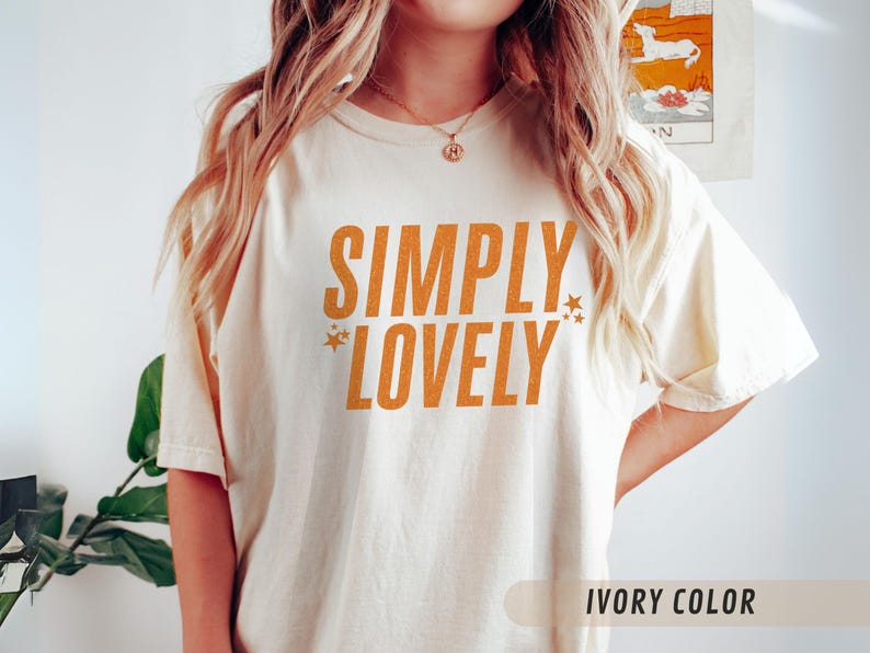 May include: Cream-coloured t-shirt featuring the orange text "SIMPLY LOVELY". The design includes star graphics. The shirt has a loose fit and short sleeves. The text "IVORY COLOR" is at the bottom.