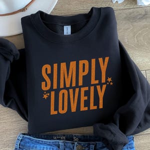 Pu&ograve; includere: Felpa nera con le parole "SIMPLY LOVELY" in lettere arancioni, con accenti a stella. La felpa &egrave; ripiegata su jeans blu e un cappello bianco.