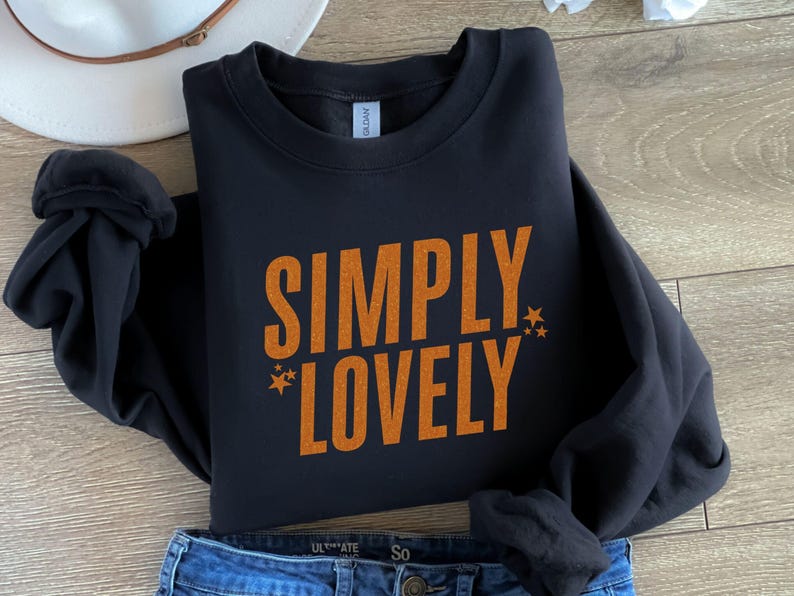 Pu&ograve; includere: Felpa nera con le parole "SIMPLY LOVELY" in lettere arancioni, con accenti a stella. La felpa &egrave; ripiegata su jeans blu e un cappello bianco.